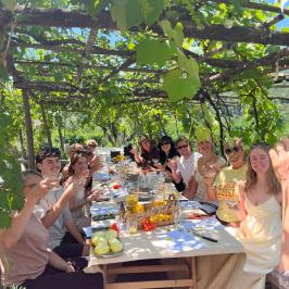 People dining outdoors under a vine-covered pergola, holding glasses, and smiling.