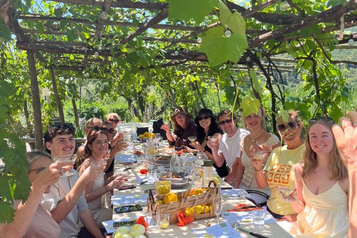 People dining outdoors under a vine-covered pergola, holding glasses, and smiling.