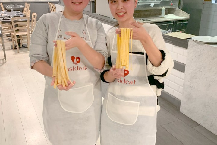 Two people in aprons holding pasta strips in a kitchen setting.