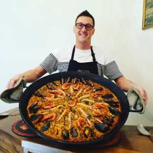 Person holding a large paella dish with seafood, wearing an apron and smiling.