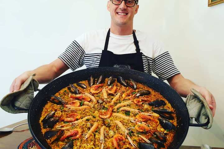 Person holding a large paella dish with seafood, wearing an apron and smiling.