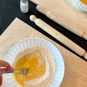 Hand mixing flour and eggs in a bowl on a wooden board with a fork.