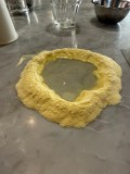Flour mound with water in center on marble countertop, glasses in the background.