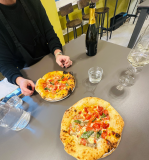 Two pizzas on a table with a bottle of wine, glasses, and a person's hands visible.