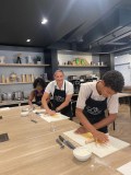 Three people in chef aprons roll dough at a kitchen counter in a cooking class.