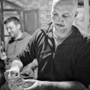 Two men in a rustic setting, one pouring liquid from a bottle, barrels visible in the background.