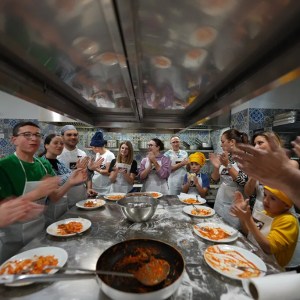 People in a kitchen wearing aprons, clapping around a table with plates of food.