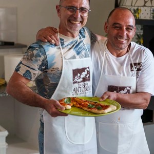 Two smiling men wearing aprons holding a plate of pizza slices.