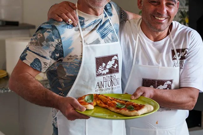 Two smiling men wearing aprons holding a plate of pizza slices.