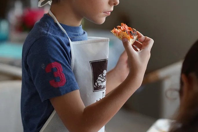 Child in yellow chef hat and apron eating a slice of pizza.