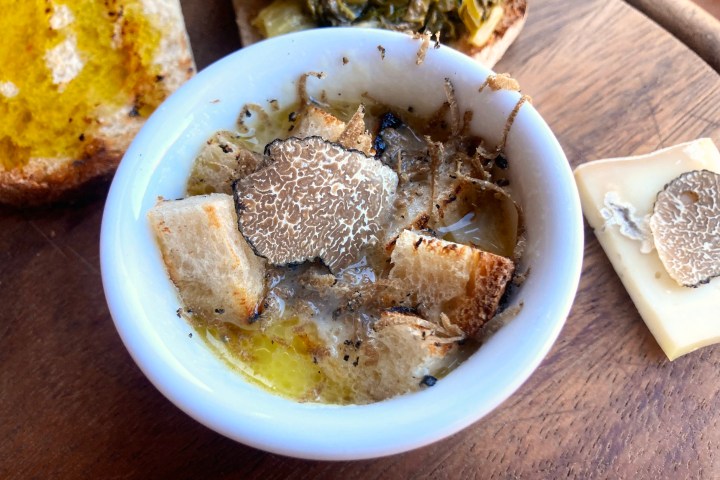 Bowl of soup with croutons and truffle slices, with cheese and bread on wooden board.