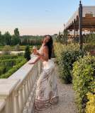 Woman in floral dress holds wine glass on terrace with scenic garden view.