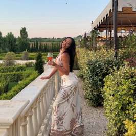 Woman in floral dress holds wine glass on terrace with scenic garden view.
