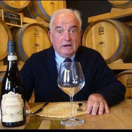 Man sitting at a table with a wine bottle and glass, wine barrels in the background.