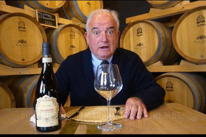 Man sitting at a table with a wine bottle and glass, wine barrels in the background.