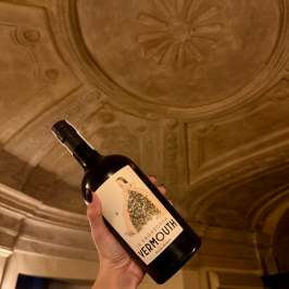 Hand holding a vermouth bottle under an ornate ceiling.