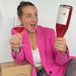 Person in pink blazer holding wine glass and bottle, sitting on cardboard box.