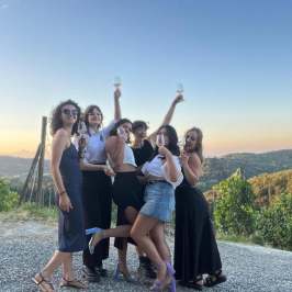 Group of six women celebrating outdoors at sunset with drinks.