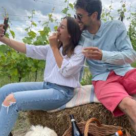 Couple taking a selfie on hay bale in vineyard, dog and picnic basket nearby.