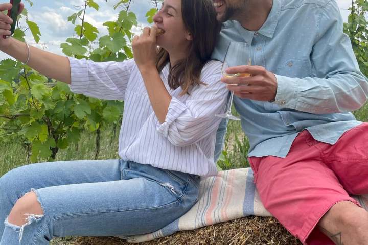Couple taking a selfie on hay bale in vineyard, dog and picnic basket nearby.