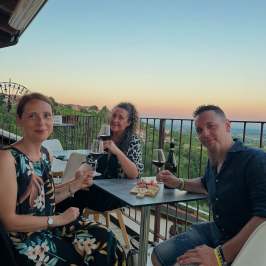 Three people sitting at a balcony table, holding drinks, with a scenic view at sunset.