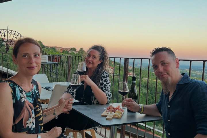 Three people sitting at a balcony table, holding drinks, with a scenic view at sunset.