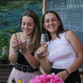 Two smiling women outdoors, one holding a glass, the other writing. Pink flowers and chalkboard in view.