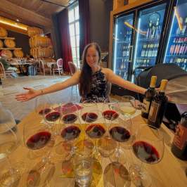 Smiling woman in a wine cellar with multiple glasses of red wine.