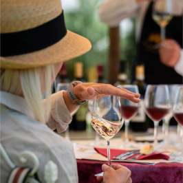 Person with hat holding wine glass at a table with more wine glasses.