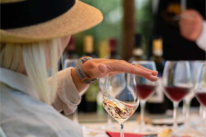 Person with hat holding wine glass at a table with more wine glasses.