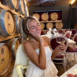 Woman in white dress holding wine glass, seated by wooden barrels.