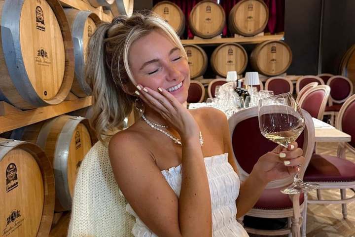 Woman in white dress holding wine glass, seated by wooden barrels.