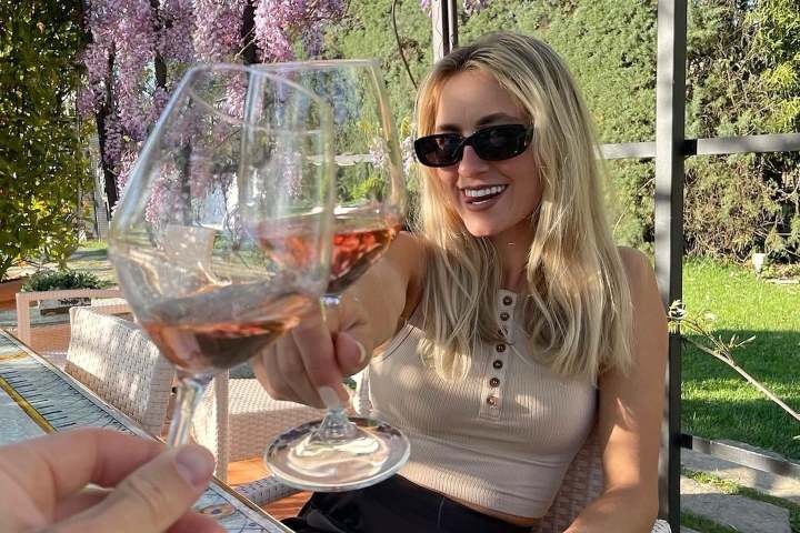 Woman sitting outdoors clinking wine glasses with a person, surrounded by flowers.