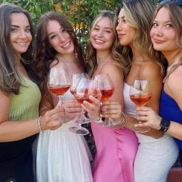 Five women smiling and holding glasses of rose wine outdoors.
