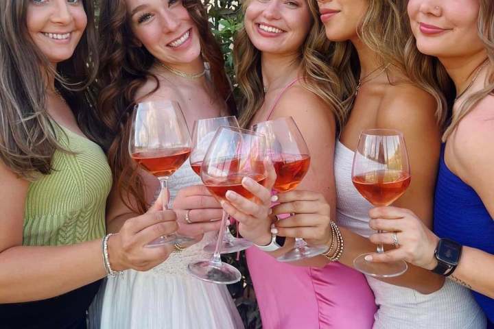 Five women smiling and holding glasses of rose wine outdoors.