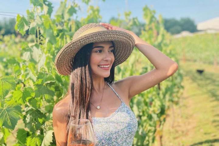 Woman in sundress and hat holding wine glass in vineyard on a sunny day.