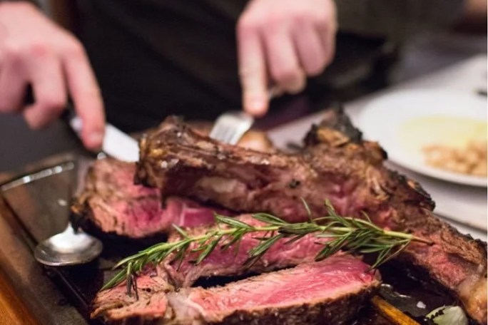Person slicing juicy grilled steak garnished with rosemary on a wooden board.