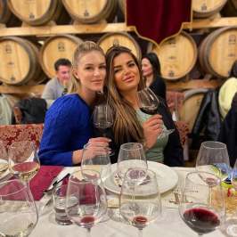 Two women sitting at table with wine glasses in a winery setting.