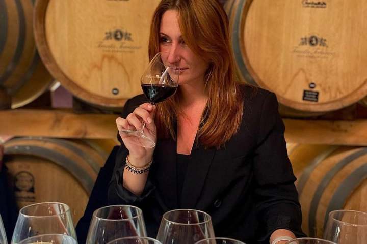 Woman tasting red wine among barrels and glasses.
