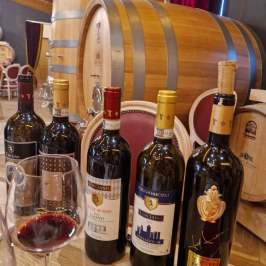 Five wine bottles and a glass of red wine in a winery with large barrels.