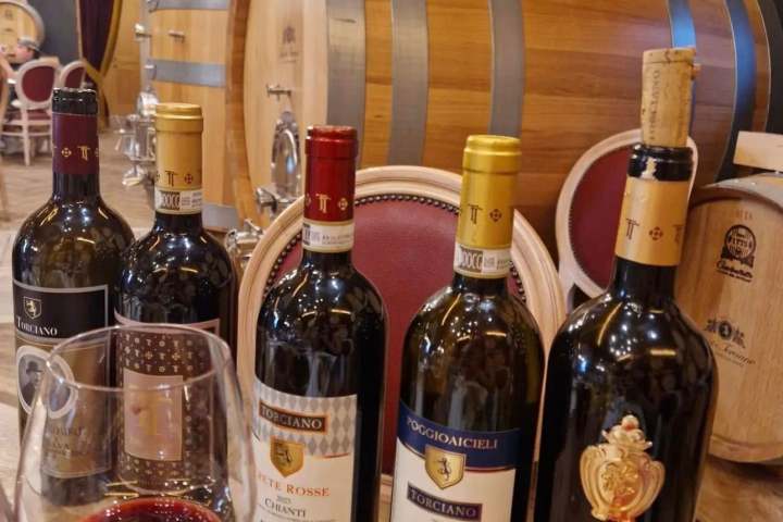 Five wine bottles and a glass of red wine in a winery with large barrels.