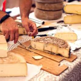 Person cutting cheese on wooden board at a market stall.