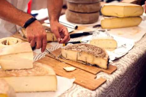 Person cutting cheese on wooden board at a market stall.