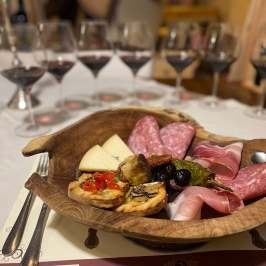 Wooden platter with meats, cheese, and bread. Glasses of red wine in the background.