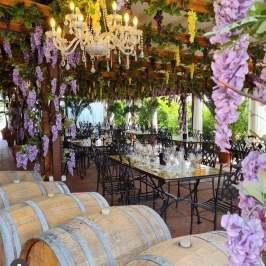 Outdoor dining area with purple flowers, chandelier, wooden barrels, and black chairs.