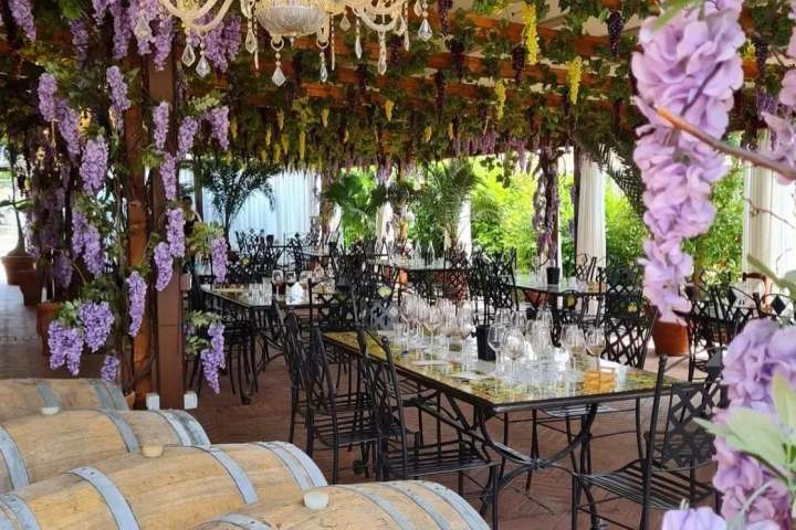 Outdoor dining area with purple flowers, chandelier, wooden barrels, and black chairs.