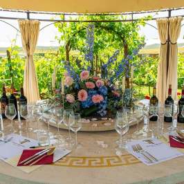 Outdoor table set with wine glasses, bottles, and a floral centerpiece under a pergola in a vineyard setting.