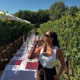 Woman drinks wine by a long outdoor table in a vineyard.