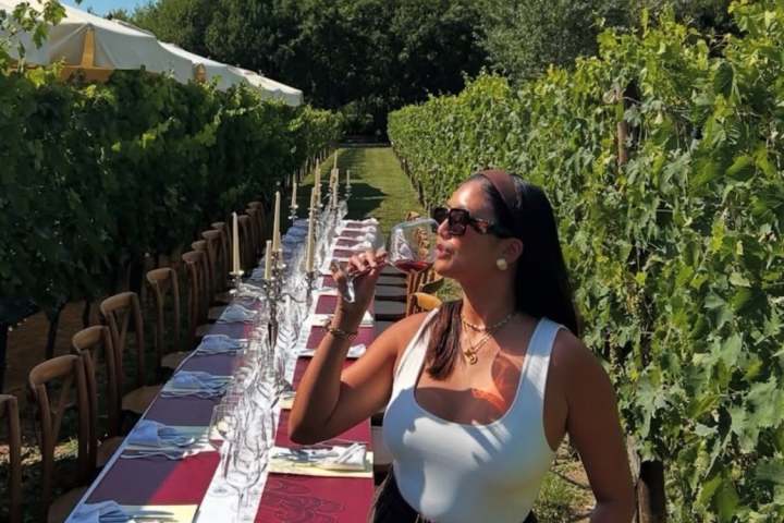 Woman drinks wine by a long outdoor table in a vineyard.