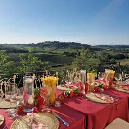 Outdoor dining table with red tablecloth, pasta, and wine, overlooking vineyard landscape.
